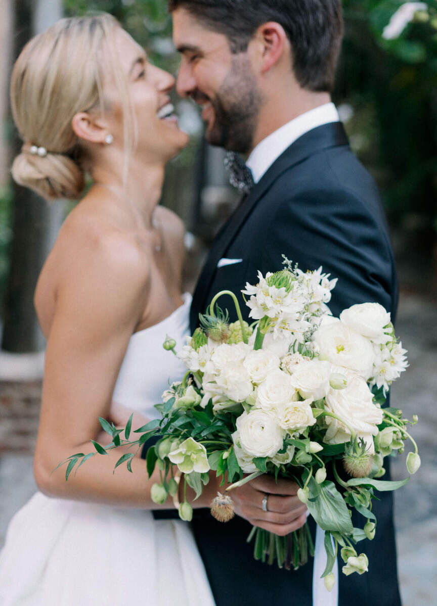 A bride, wearing a twisted, low updo and holding a bouquet made of white blooms smiles at her groom at their destination wedding.