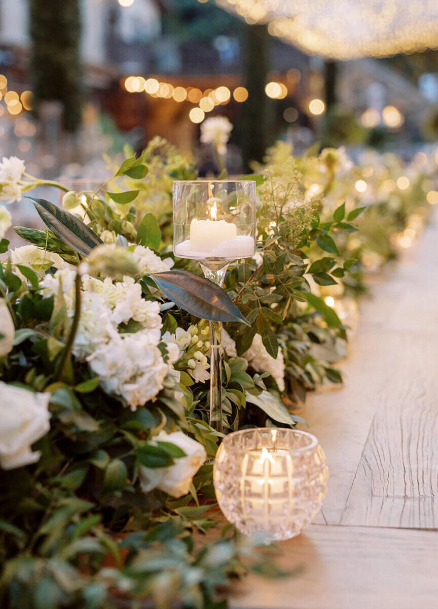 Rectangular tables at a destination wedding reception were set with long arrangements of greenery, white flowers, and candles in a mixture of vessels.