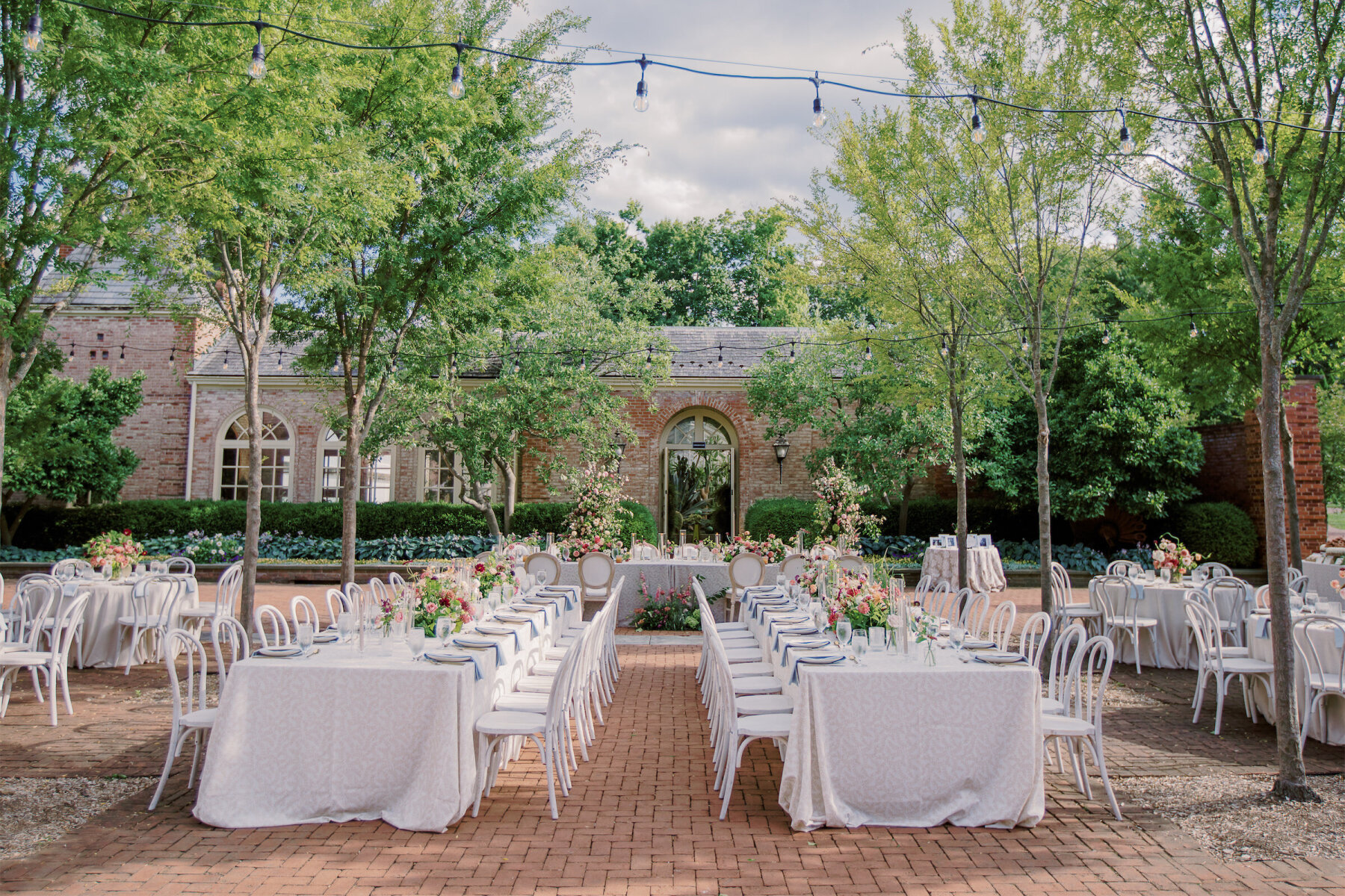 An enchanted garden wedding reception, with a mix of long and round tables, set under strands of bistro bulb lighting.
