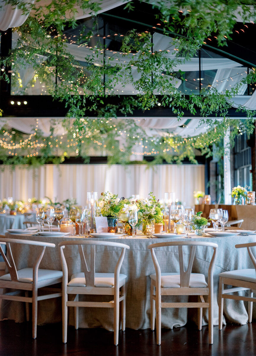 Pale wood wishbone chairs surround oval reception tables, set under a canopy of draping, twinkle lights, and greenery at a enchanted waterfront wedding in Chicago.