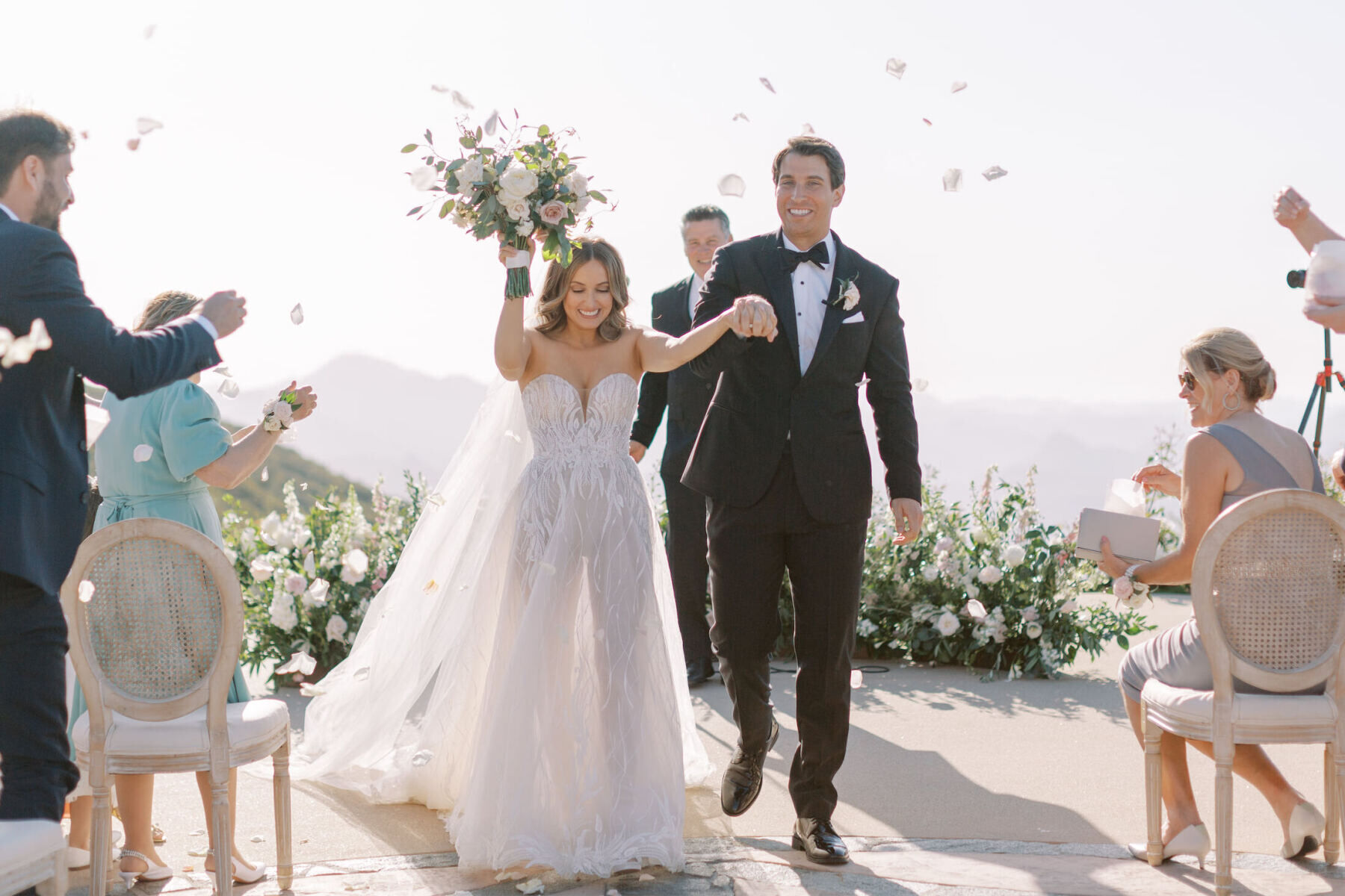 A bride and groom couple recess up the aisle at their estate wedding as guests toss rose petals in the air.