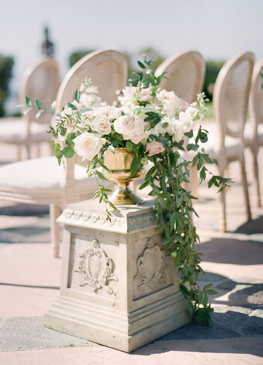 Upholstered chairs and an ornate pedestal under a gold urn of flowers create a beautiful wedding ceremony environment.