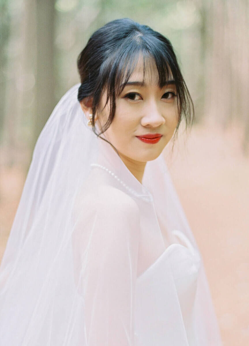 A bride with curtain bangs and wearing a veil and red lipstick looks at the camera for a portrait at her forest wedding.