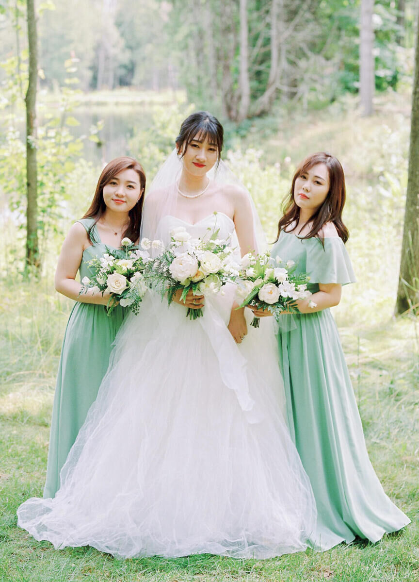 A bride and her two bridesmaids (who wear soft green dresses) pose for a group portrait at a forest wedding, where the flowers were all-white and green.
