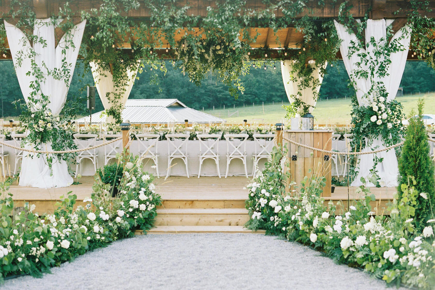 A flower-lined pathway to an outdoor wedding reception on a deck decorated with hanging greenery, drawpery, and linen-covered tables.