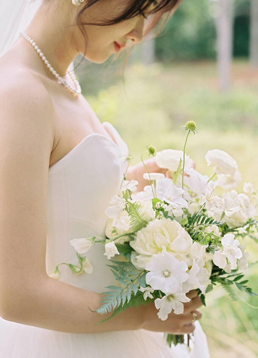 A bride holds her bouquet—made of all whit flowers and fresh ferns—at her forest wedding in Canada.