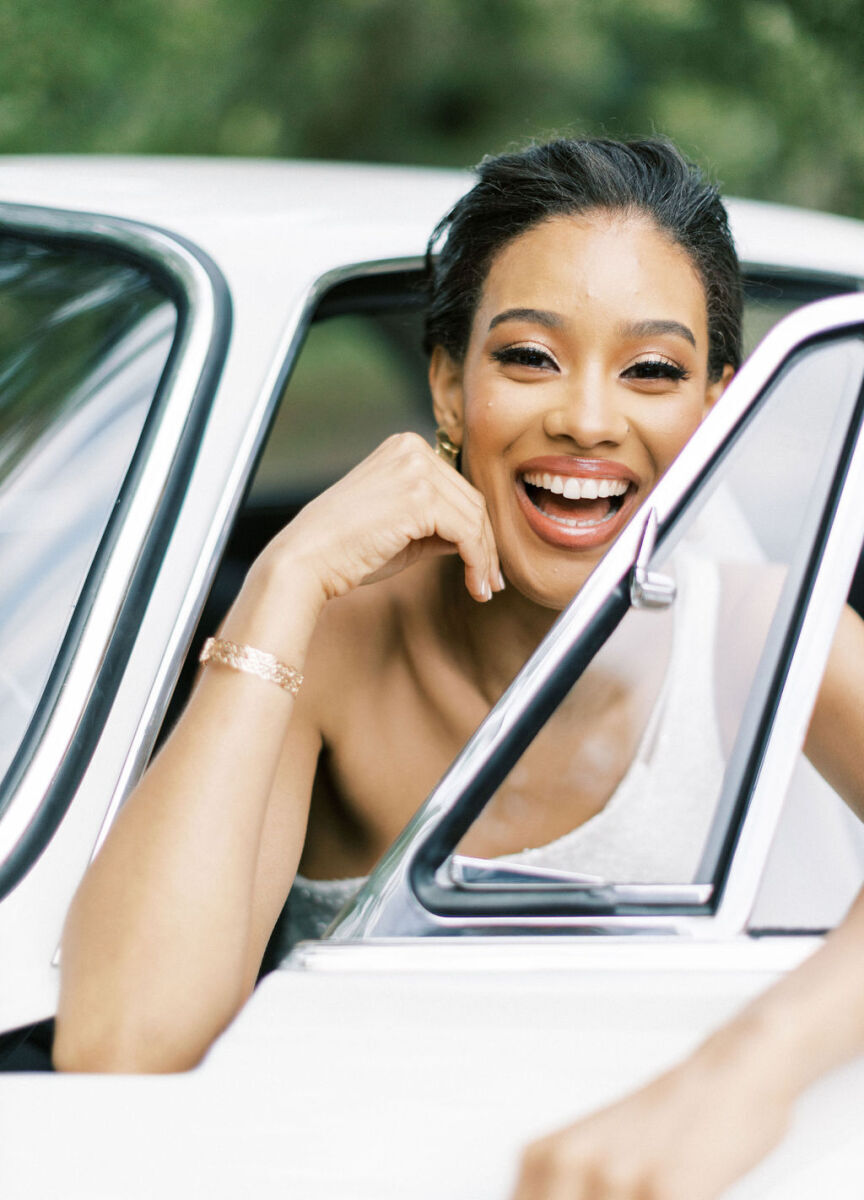 A smiling bride wearing gold jewelry and a loose updo, in a classic car at her French wedding.