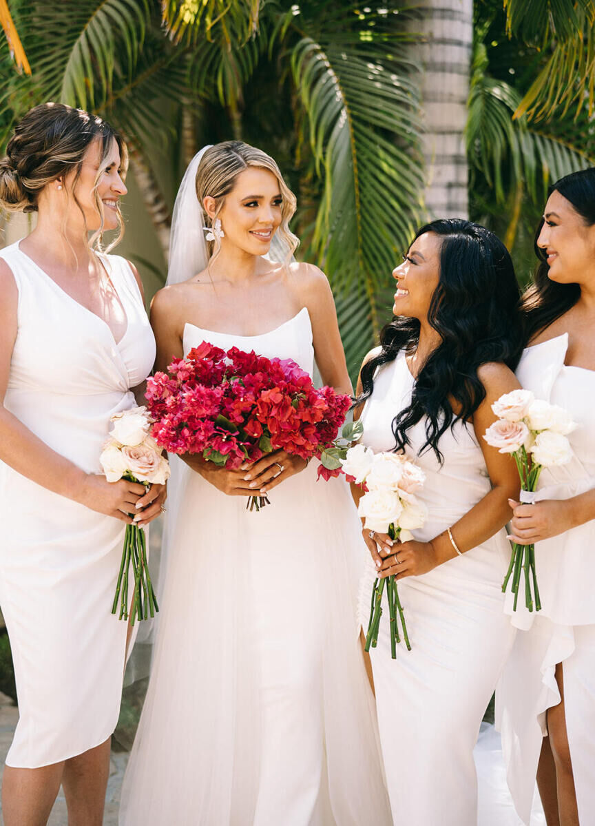 A bride and her bridesmaids all wear white and hold bouquets of singular flower types ahead of her glam beach wedding ceremony at Esperanza.