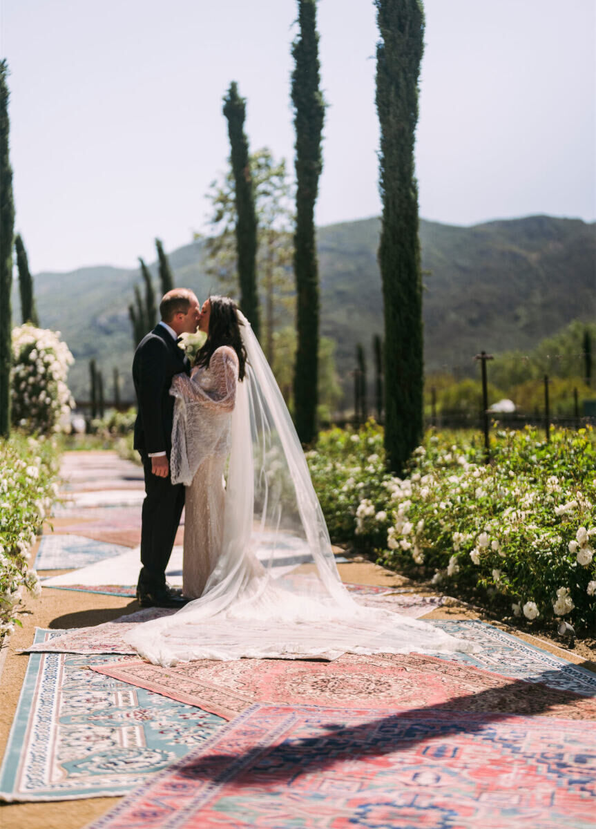 A groom kisses a bride on a pathway of overlapping rugs at their glam enchanted wedding.
