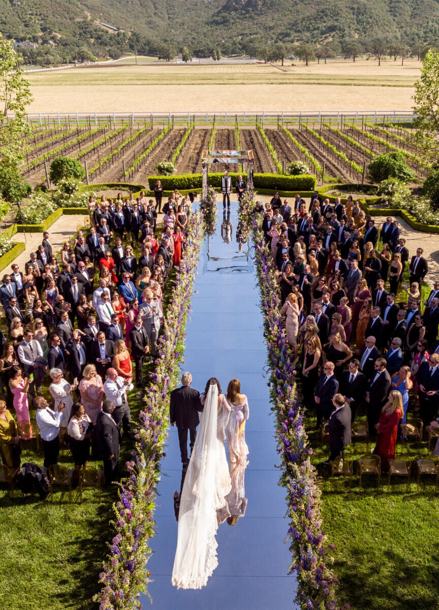 A bride and her parents proceed down a mirror-lined ceremony aisle at her glam enchanted wedding on a vineyard.