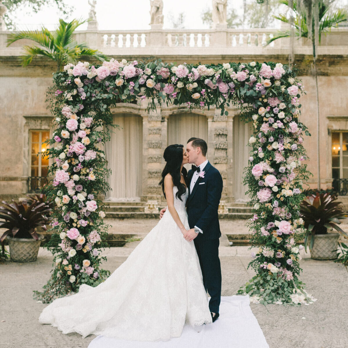 A bride and groom kiss in front of their floral arch during their glam, garden wedding.