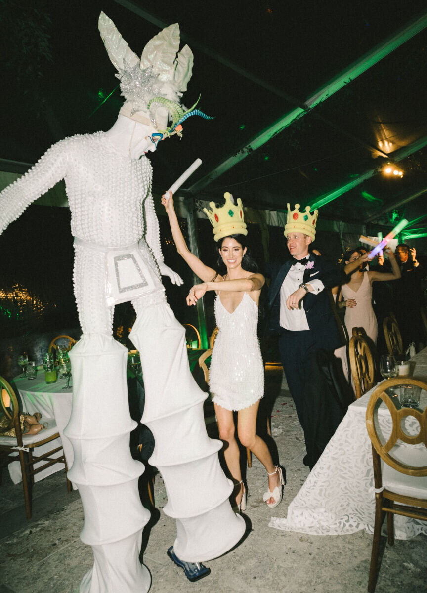 A stilt walker leads a bride and groom through their reception space during an hora loca at their glam, garden wedding.