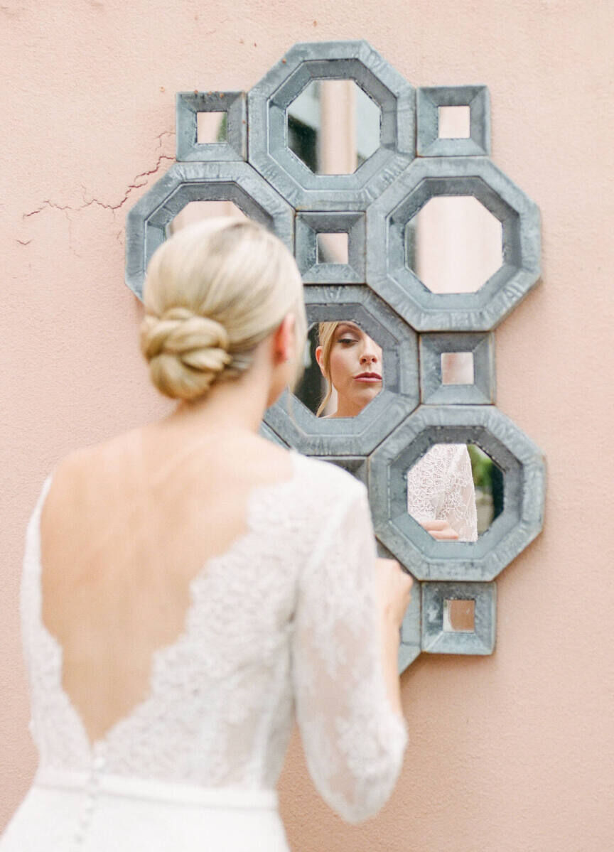 A bride (wearing a knotted, low updo) checks her makeup in a mirror during her Glenmere Mansion wedding.