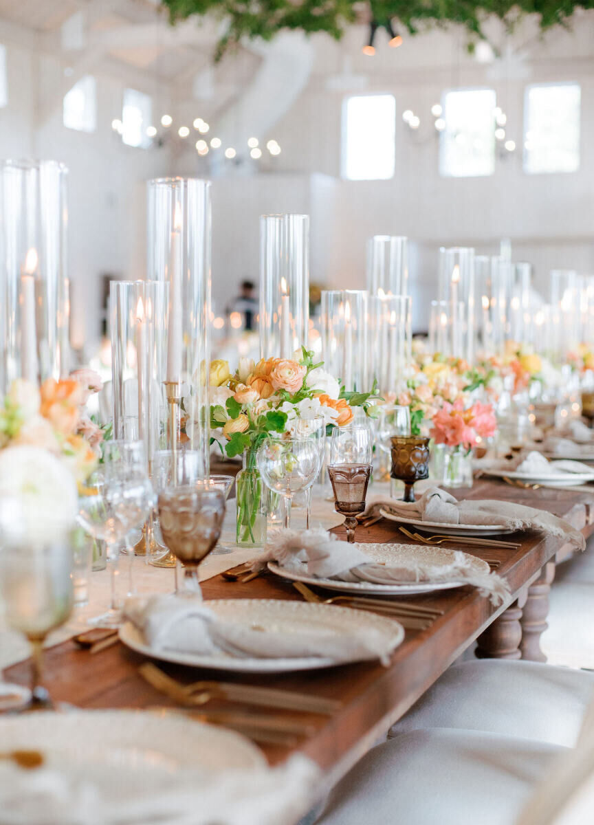 A long table set inside the barn at a Glenmere Mansion wedding reception incorporated vintage glassware, peach and white centerpieces, and knotted linen dinner napkins.