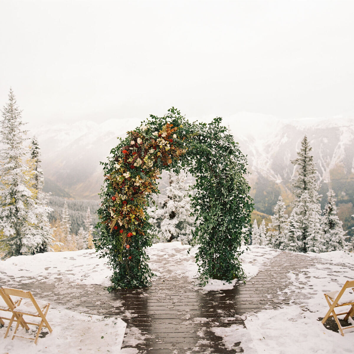 Holiday Wedding: An outdoor snowy wedding ceremony set-up with pine trees in the background. There's a wedding arch adorned with greenery and red florals.