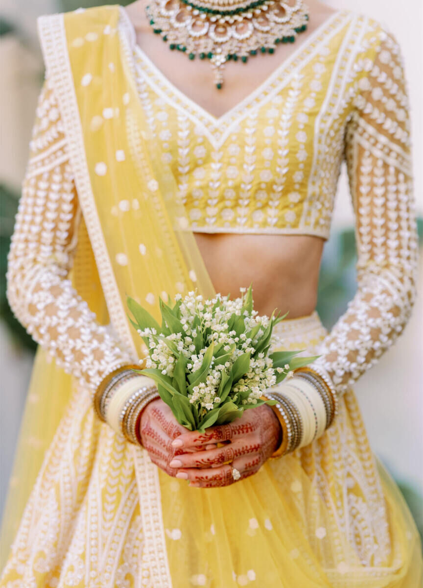 A bride, wearing a yellow lehenga, holds a petite bouquet of lily of the valley at her Indian fusion wedding.