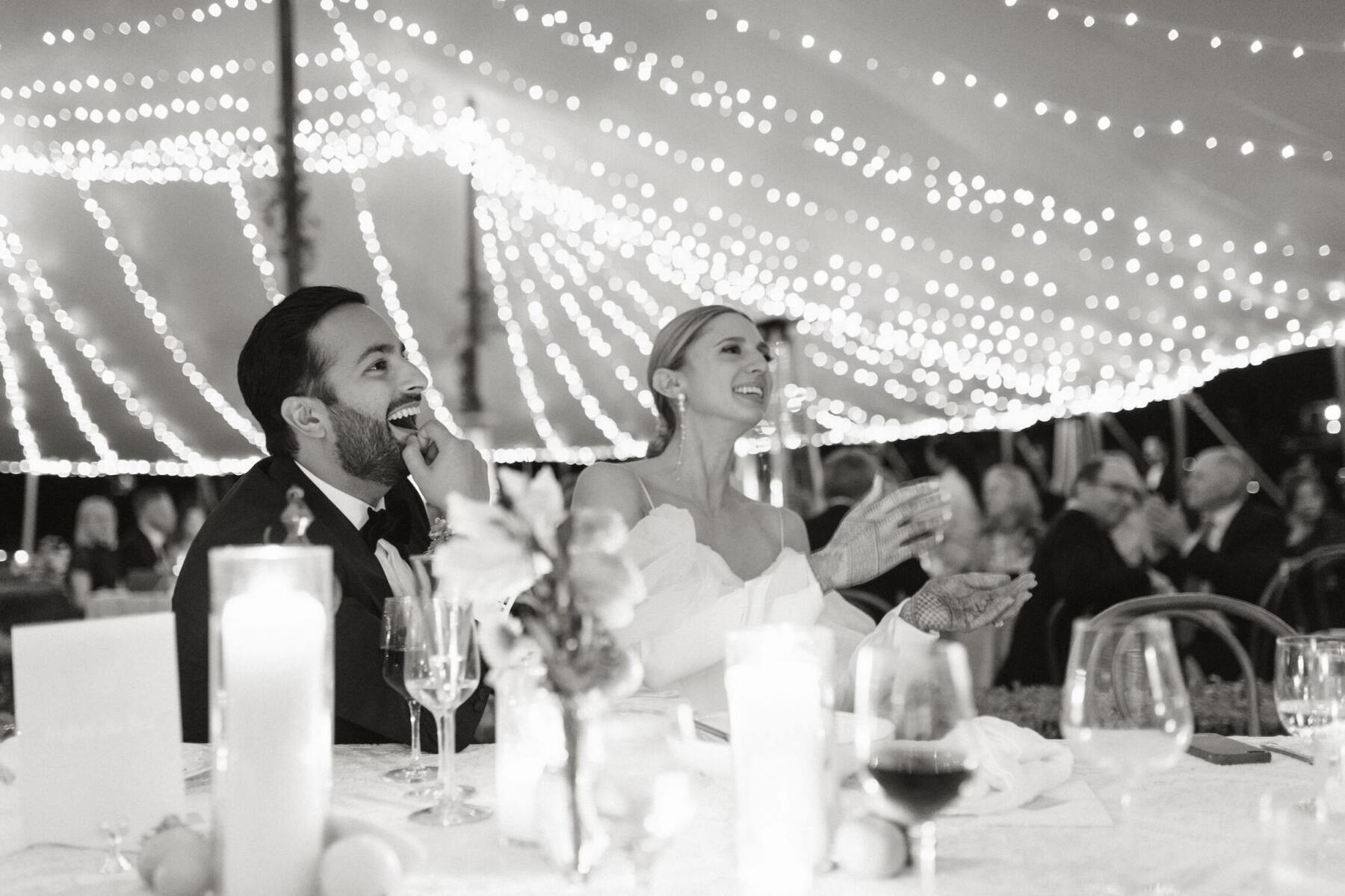 A groom and bride laugh during their Indian fusion wedding reception which took place in a tent with lights strung overhead.