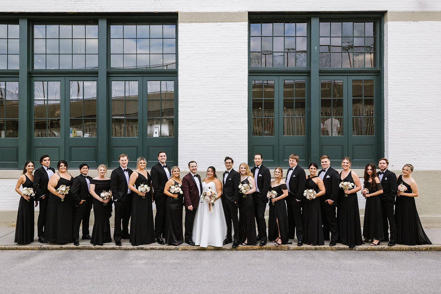A wedding party poses for a group portrait during an industrial wedding in Baltimore. Bridemaids wore black gowns, groomsmen wore black tuxedos, and the groom wore a burgundy dinner jacket. 