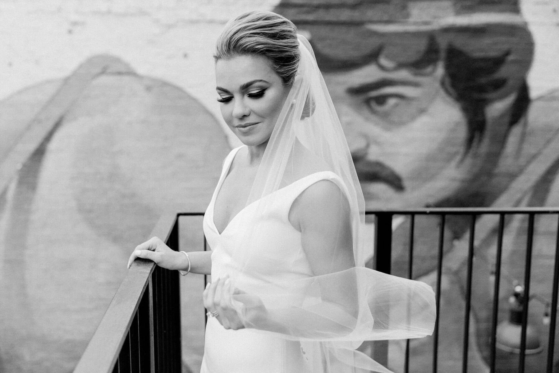 A bride with lush lashes and a sleek updo and veil poses for a portrait in front of a mural.