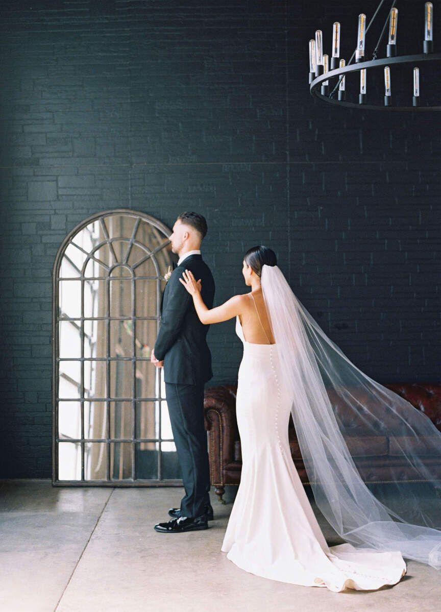 Industrial Wedding Venues: A bride tapping a groom on the shoulder for a first look. They're standing in front of a black brick wall with a cool window-inspired mirror and metal chandelier.