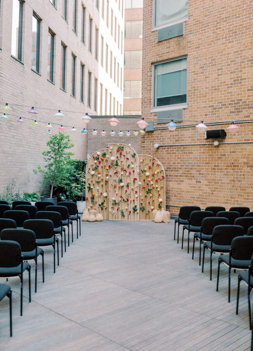 Industrial Wedding Venues: An outdoor, open-air ceremony set-up at Yours Truly DC complete with brick string lights, black seats for guests, and an edgy flower/wood wall at the front.