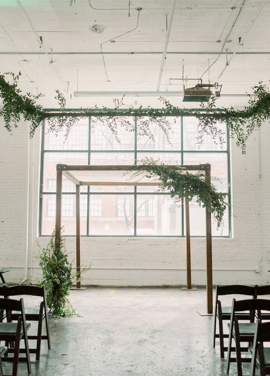 Industrial Wedding Venues: An industrial ceremony set-up with a wooden archway adorned with greenery in front of a white-brick wall and large loft-style window.
