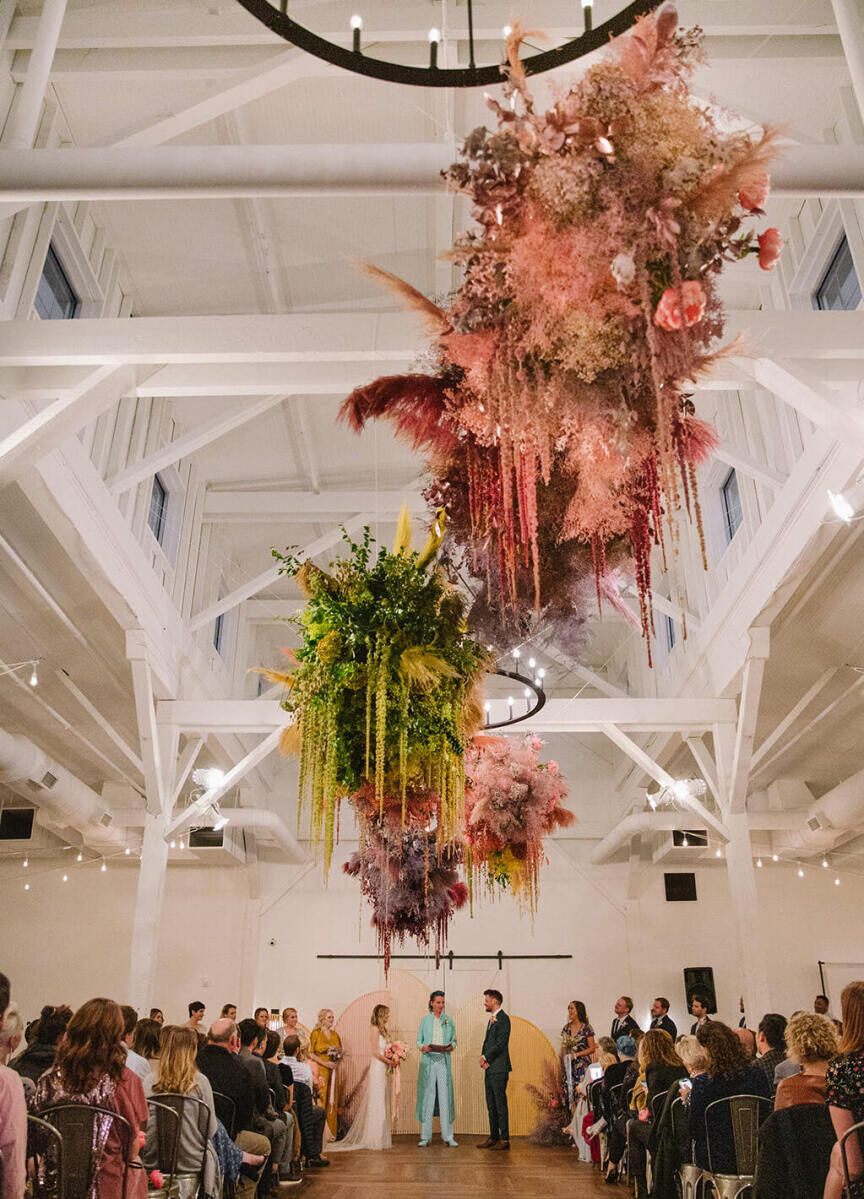Industrial Wedding Venues: Colorful floral installations hanging from the ceiling beams above a wedding ceremony at 14TENN.