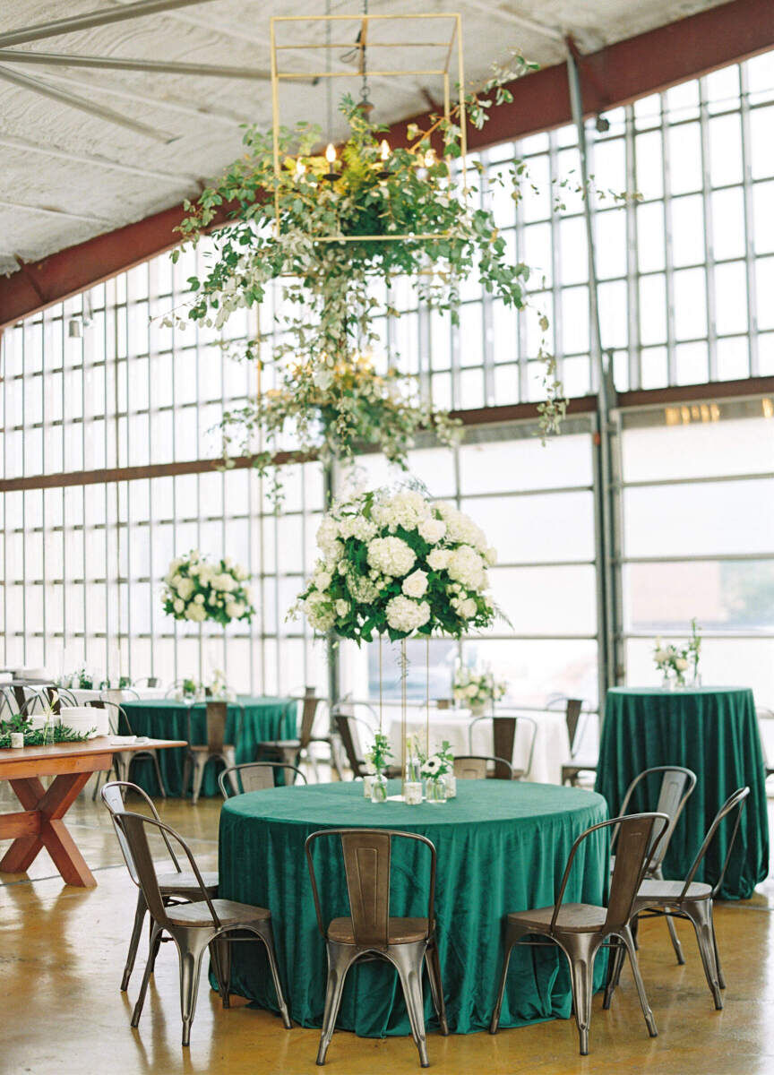 Industrial Wedding Venues: A green table with chairs sitting under hanging floral installations and in front of large greenhouse -style windows.