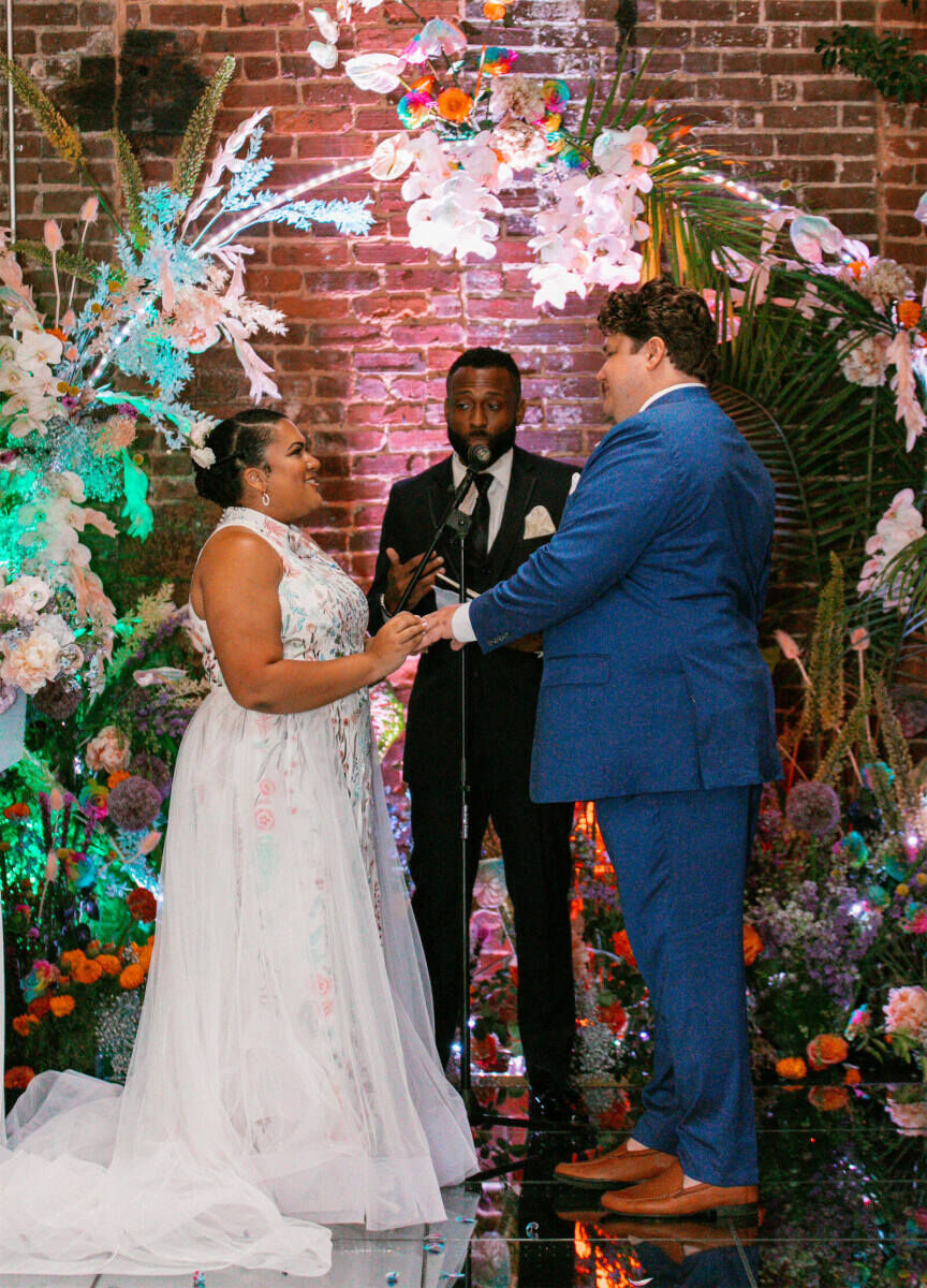 Industrial Wedding Venues: A couple getting married and holding hands while standing in front of a brick wall and circular floral installation.