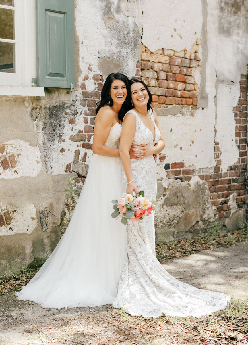 Industrial Wedding Venues: Two brides holding each other and standing close by a brick wall with turquoise shutters.