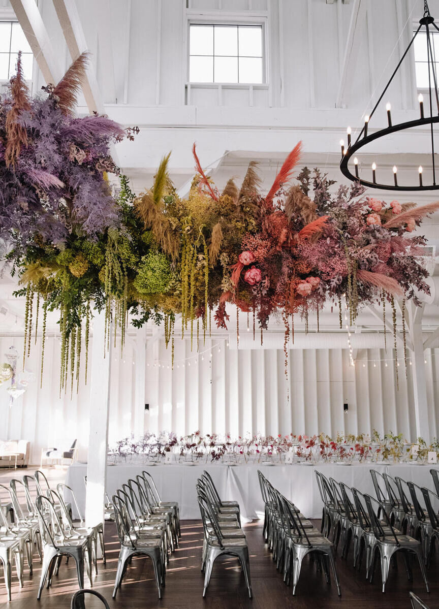 Industrial Wedding Venues: A colorful floral installation hanging above metal chairs set up for a wedding ceremony in a white interior room. A metal chandelier can be seen next to the floral installation.