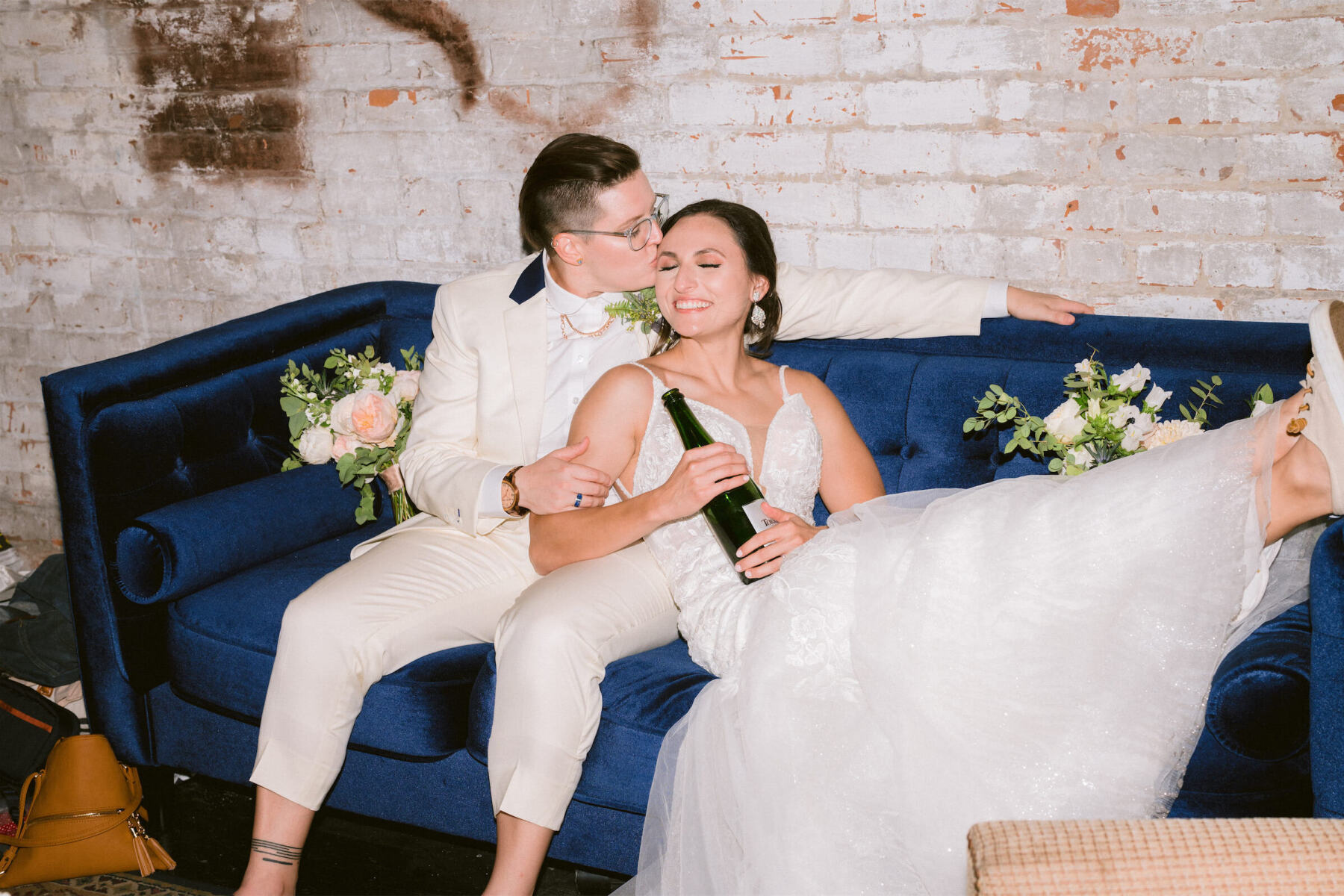 Industrial Wedding Venues: Two newlyweds snuggling on a blue couch, with one person kissing another on the cheek while the other holds a bottle of champagne and smiles.
