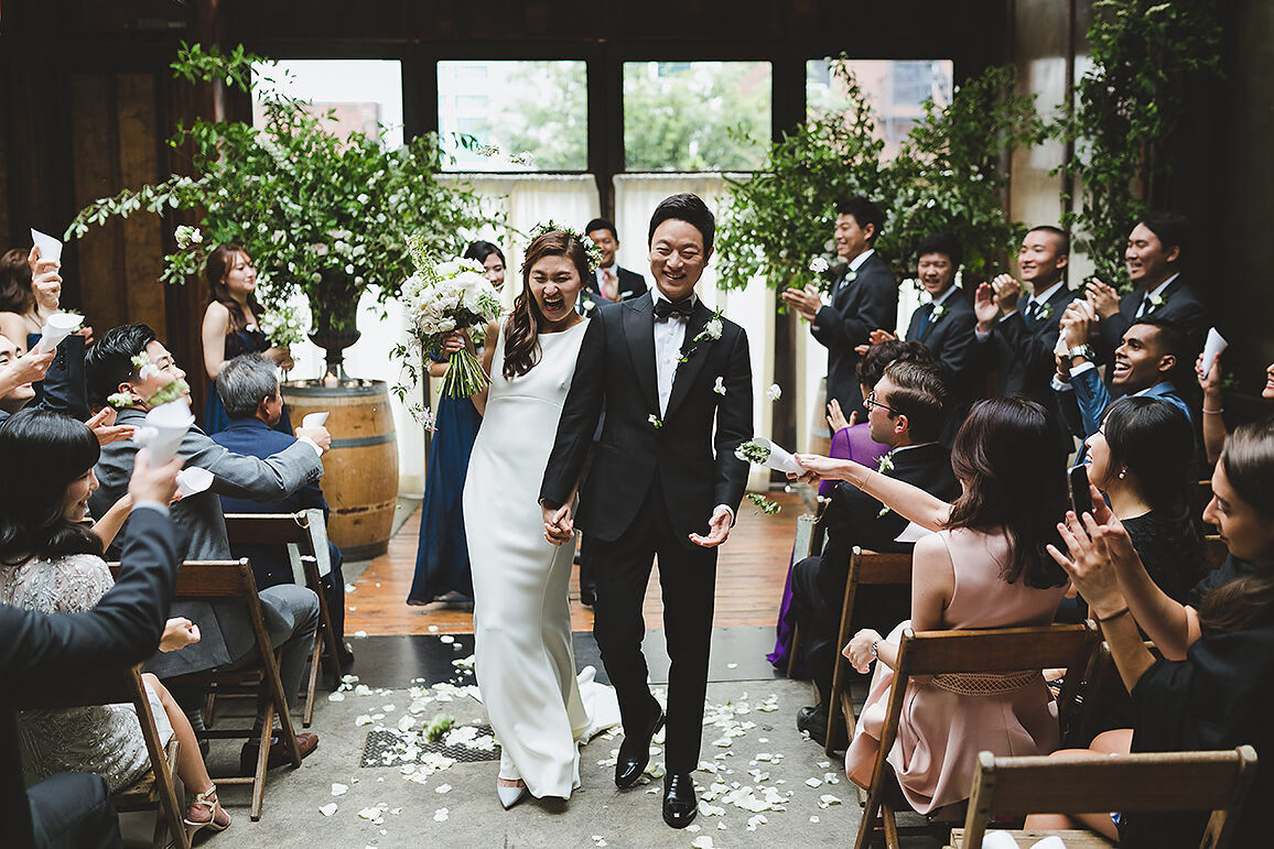 Industrial Wedding Venues: A bride and groom smiling and holding hands while walking back down the aisle together while guests toss flower petals at them at Brooklyn Winery.