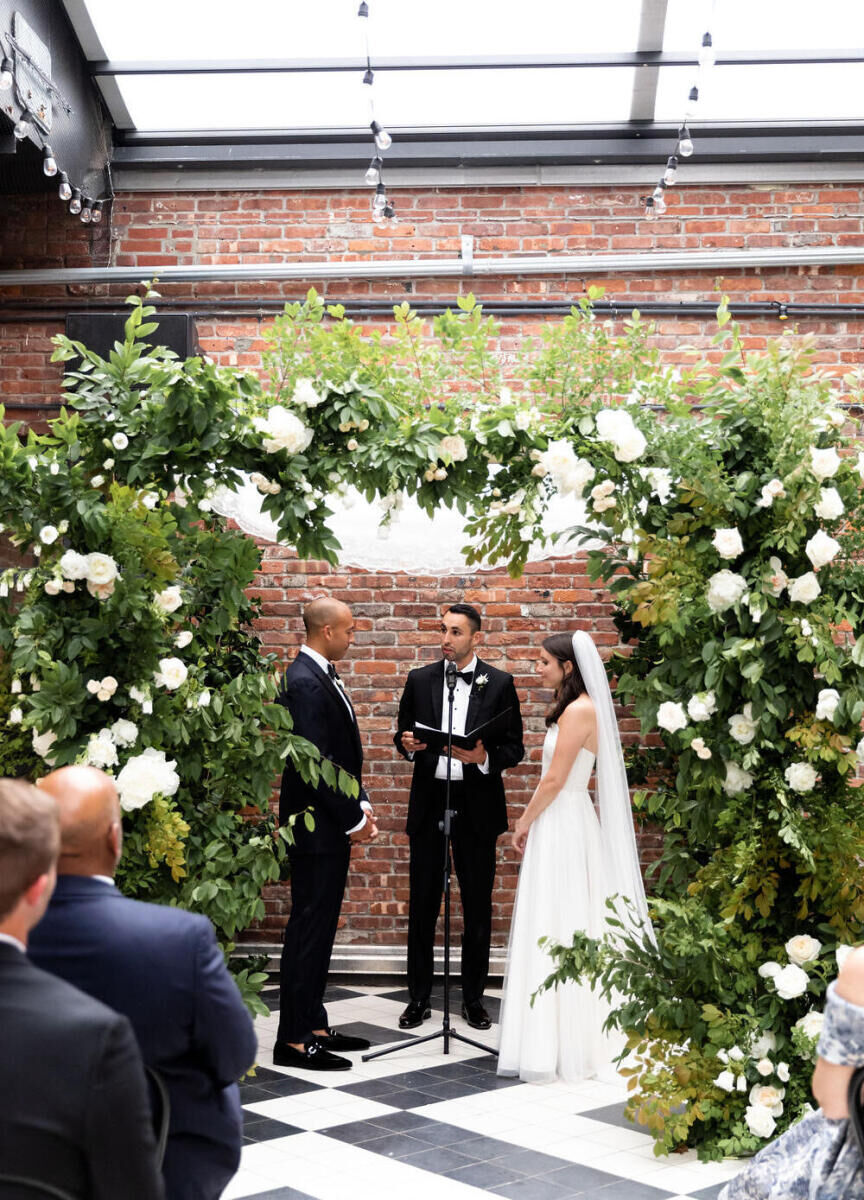 Industrial Wedding Venues: A couple standing under a flower archway during their wedding ceremony at the Wythe Hotel.