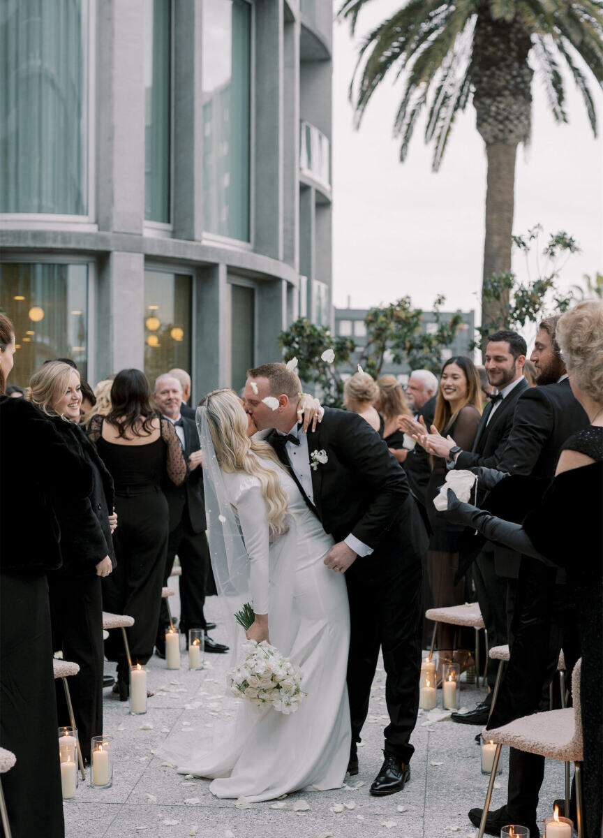 Newlyweds kiss as white rose petals are tossed at them at the end of their modern California wedding ceremony.