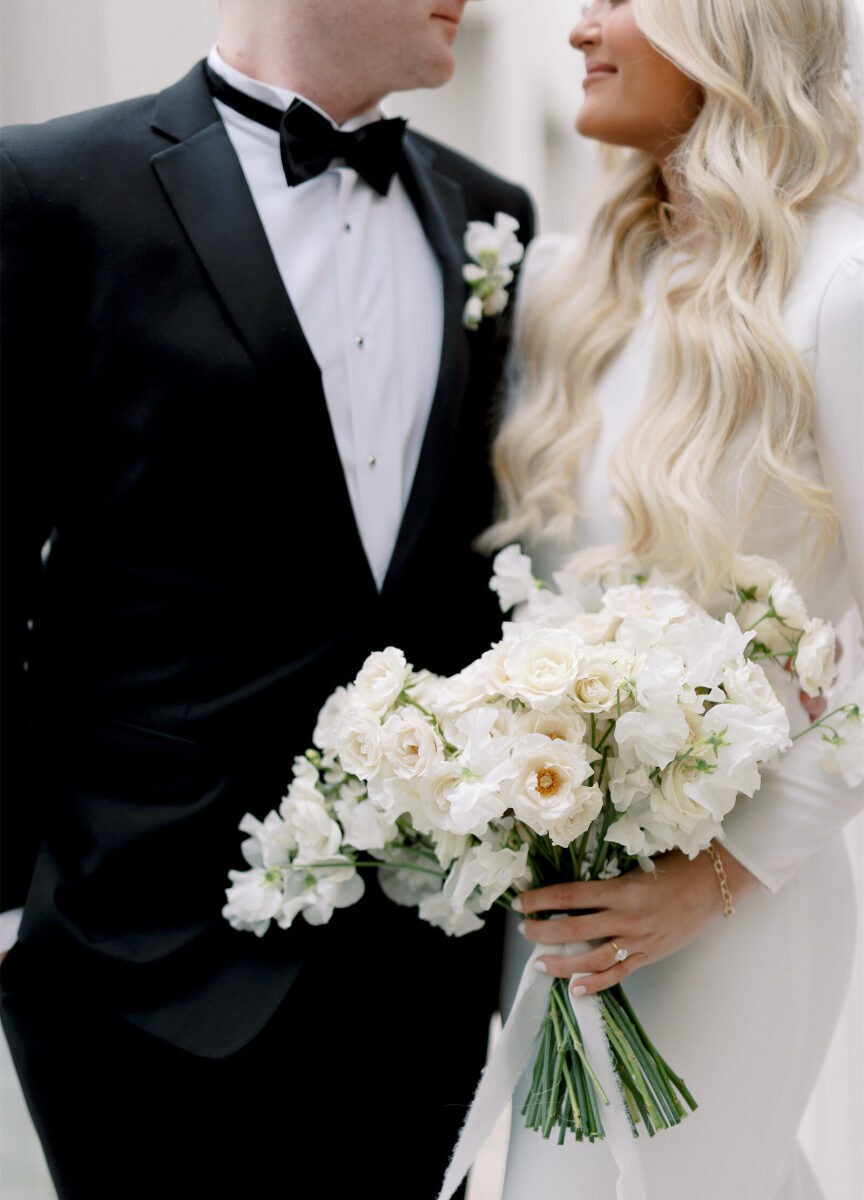 A groom and bride look at each other while she holds her all-white bouquet, which features roses and sweet peas.