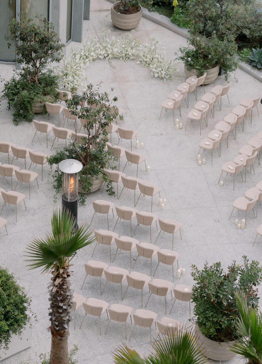 An overhead look at a modern California wedding ceremony, with textured chairs in a warm, soft taupe, set diagonally facing a low ceremony floral installation.