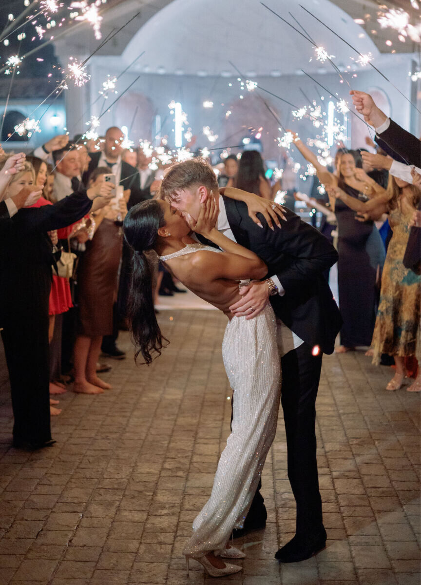 A bride wears a jumpsuit and long ponytail and kisses her now-husband as he dips her amidst their guests holding sparklers and sending them off at the conclusion of their modern fairytale wedding.