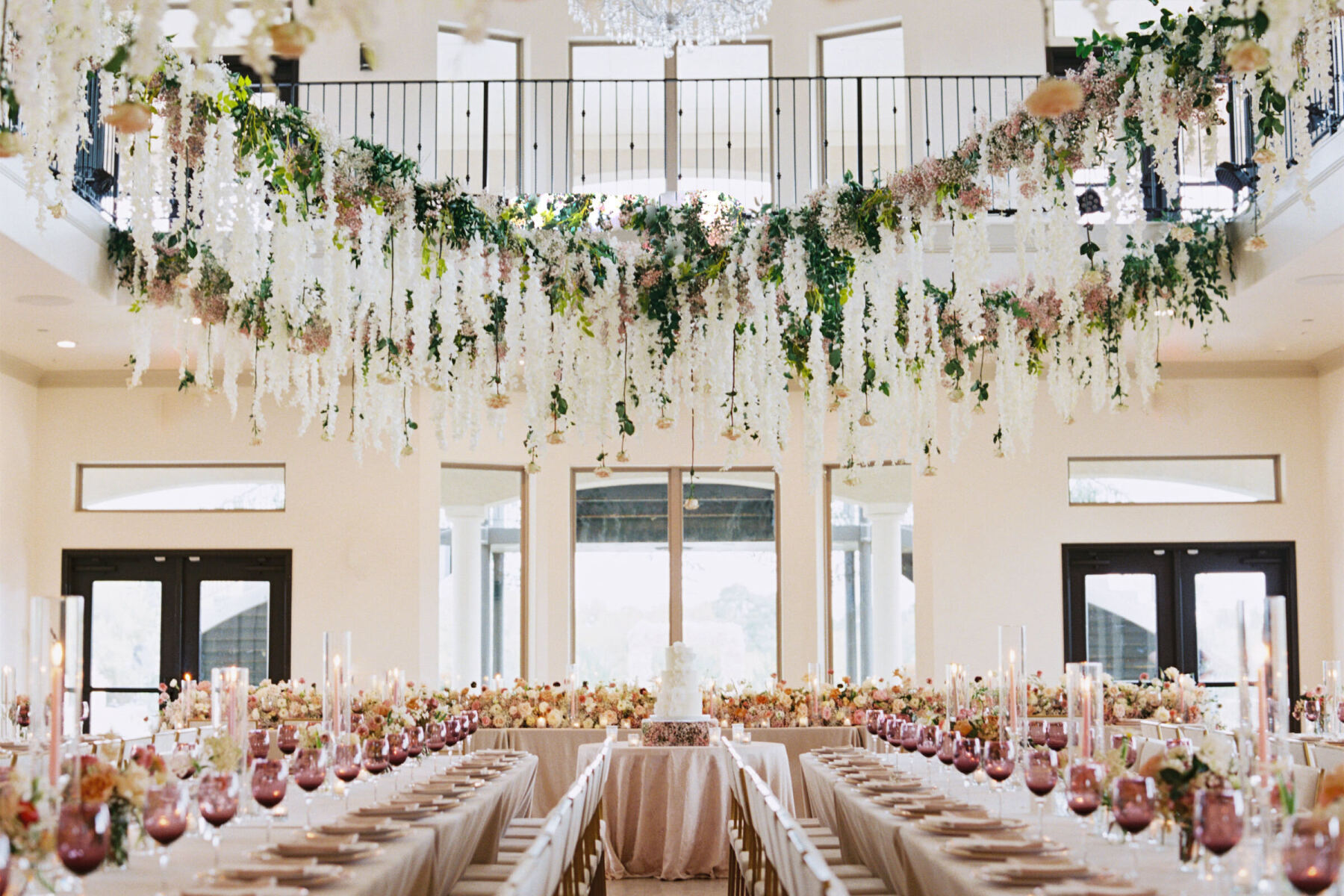 A modern fairytale wedding reception, with dripping flowers and greenery installed over a series of long reception tables decorated with plum glassware, pink taper candles, and lush centerpieces.
