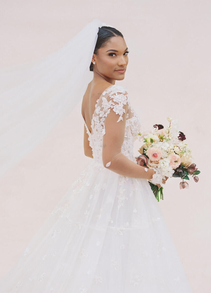 A bride wears a floral-lace adorned sleeved wedding dress and veil, and holds a white-and-pink bouquet the morning of her modern fairytale wedding.