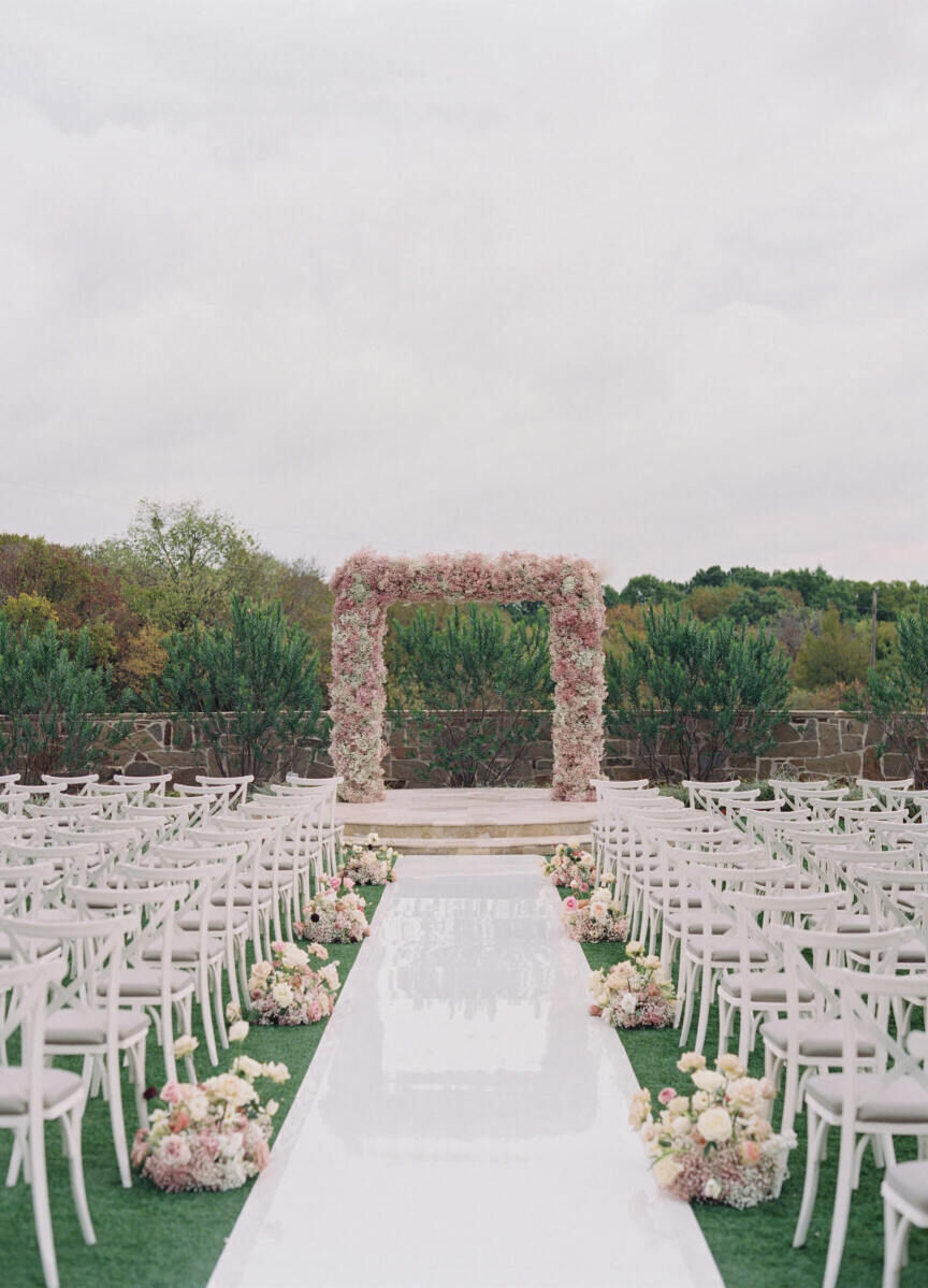A modern fairytale wedding ceremony setup with pink baby's breath arch, glossy white aisle, and white-and-pink floral arrangements.