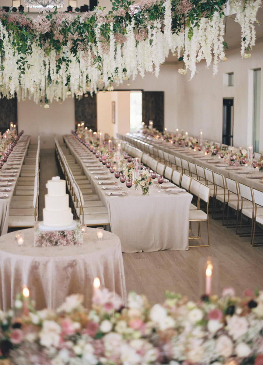 An indoor reception for a modern fairytale wedding, with long tables, flowers and greenery hanging overhead, and a cake sitting atop a clear box with pink baby's breath inside.