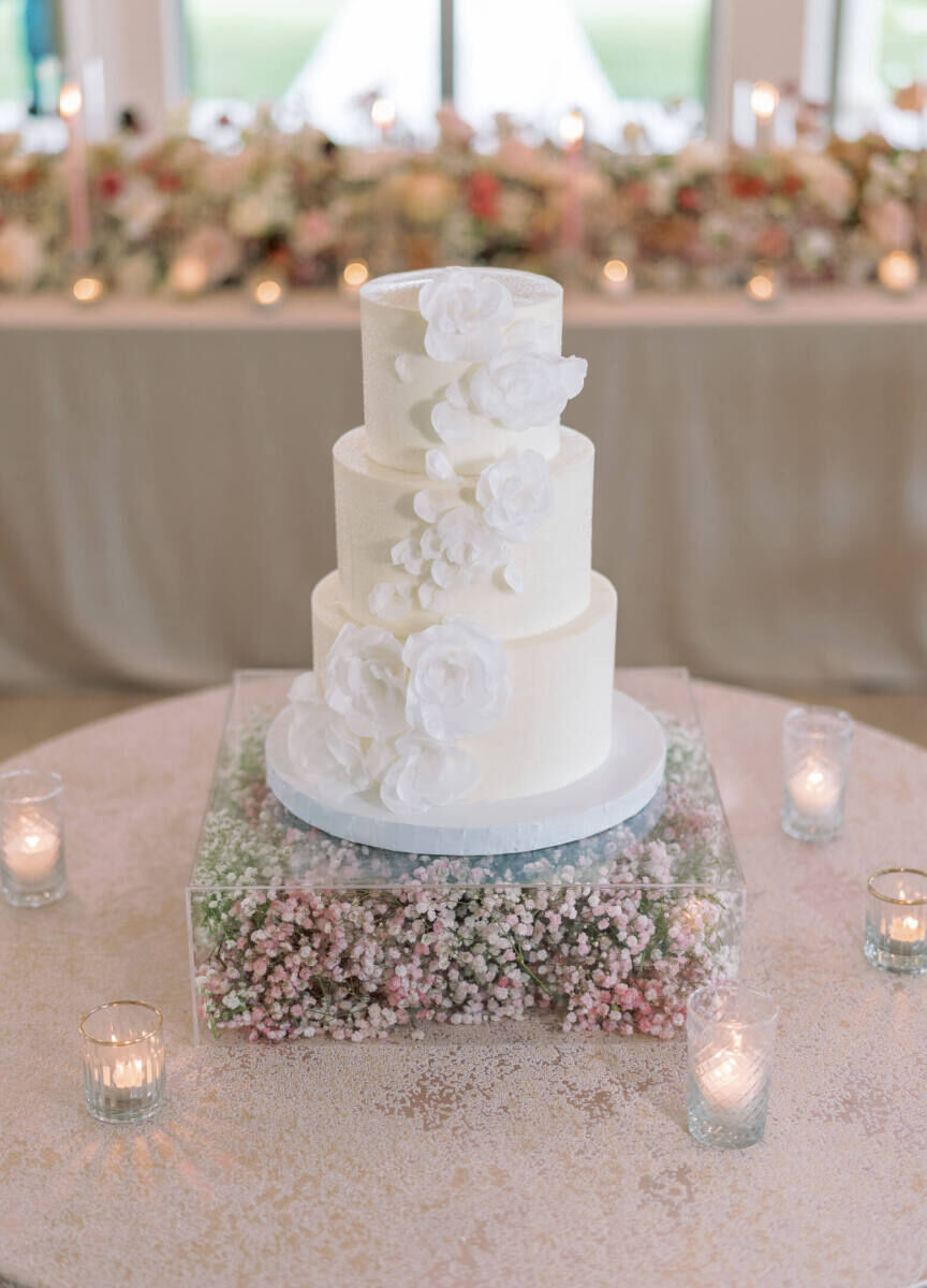 A three-tier white cake with edible white flowers cascading down the side is perched atop a clear box filled with pink baby's breath at a modern fairytale wedding.