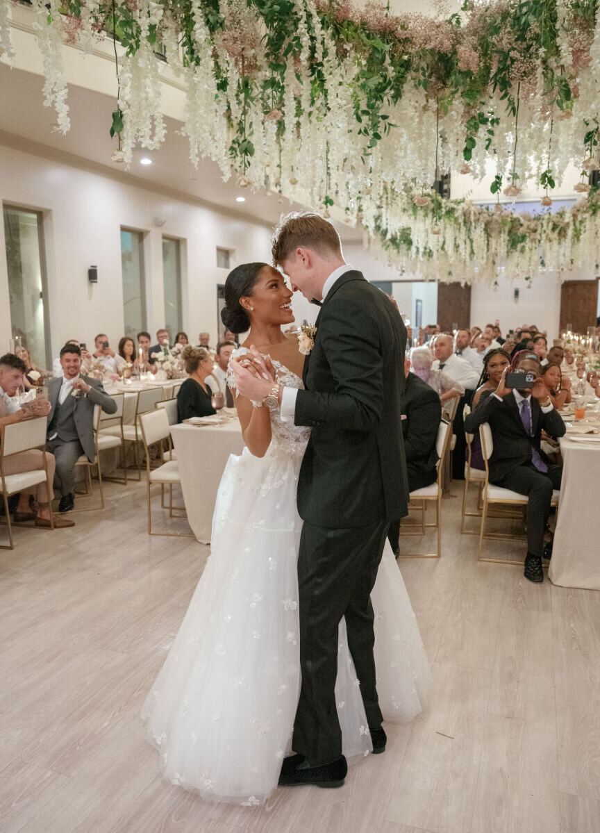 A bride and groom share a first dance under an installation of greenery and flowers at the reception of their modern fairytale wedding.