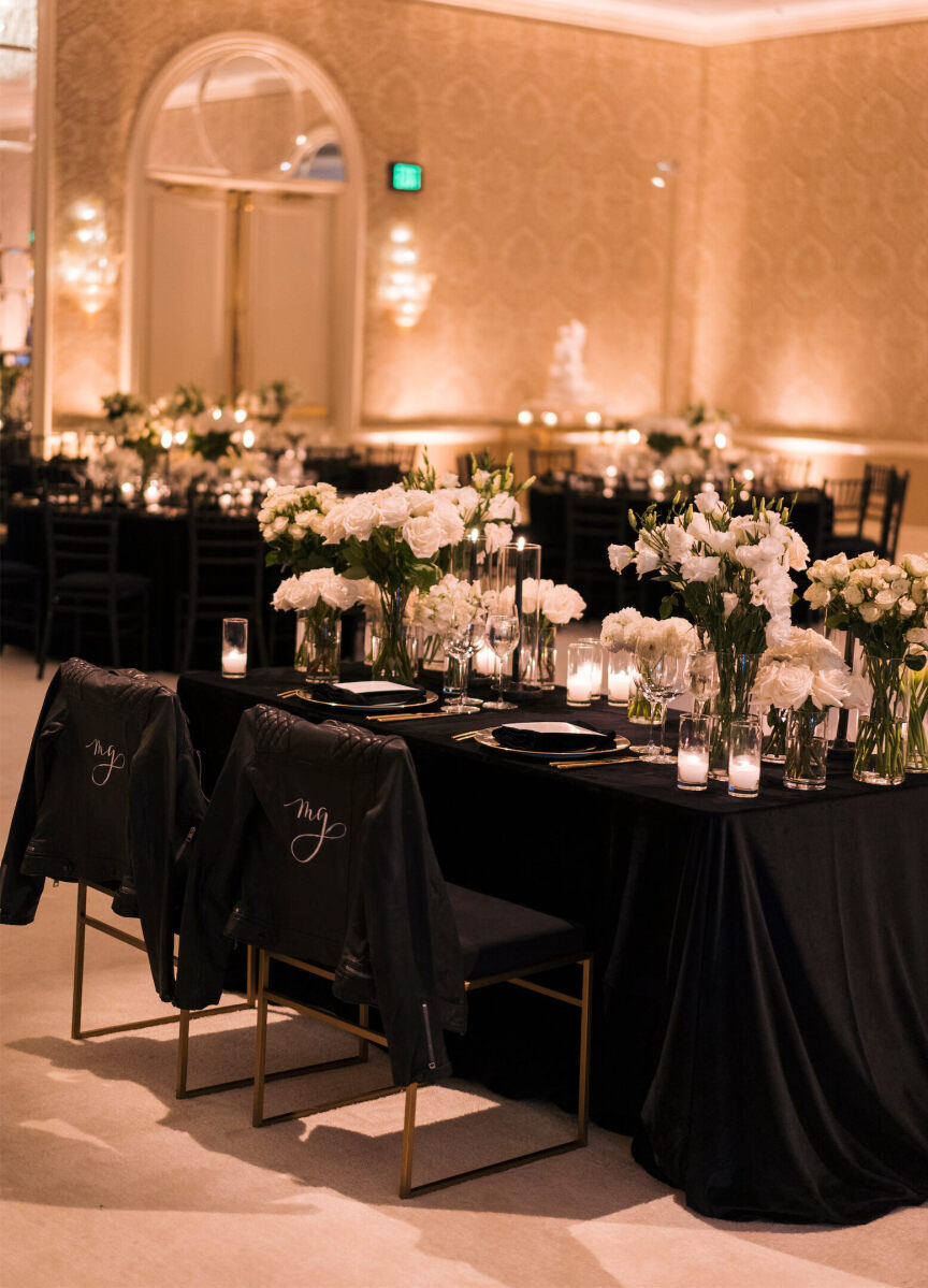 A hotel ballroom is set for a modern wedding reception using black linens and all-white flowers, with lots of candles and ambient lighting for a warm glow.