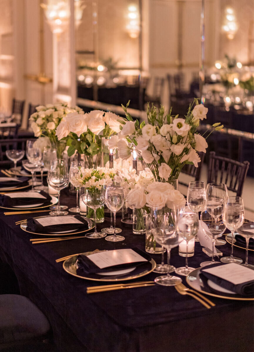A modern wedding reception in a ballroom set with black velvet linens, all-white centerpieces, and accents of gold on the tabletop.