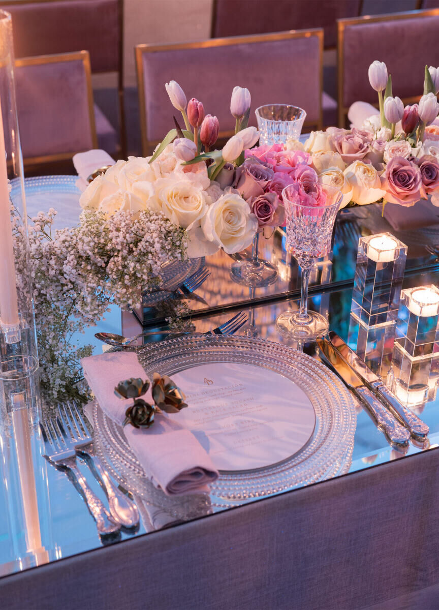 Clear chargers and candle holders reflect on a mirrored table, set with a 3-D napkin ring and clusters of pink and white flowers.