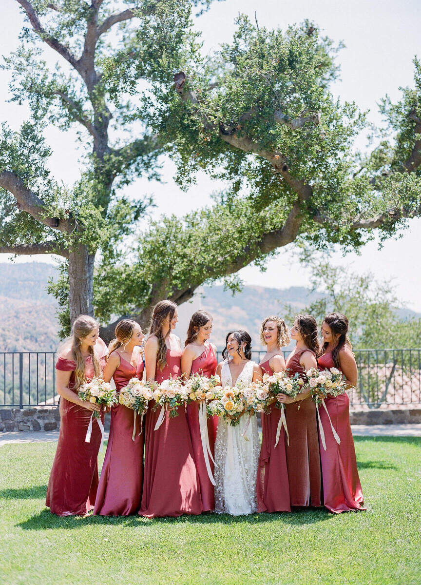 A bride is surrounded by her bridesmaids in their berry-hued dresses, all holding bouquets and smiling.