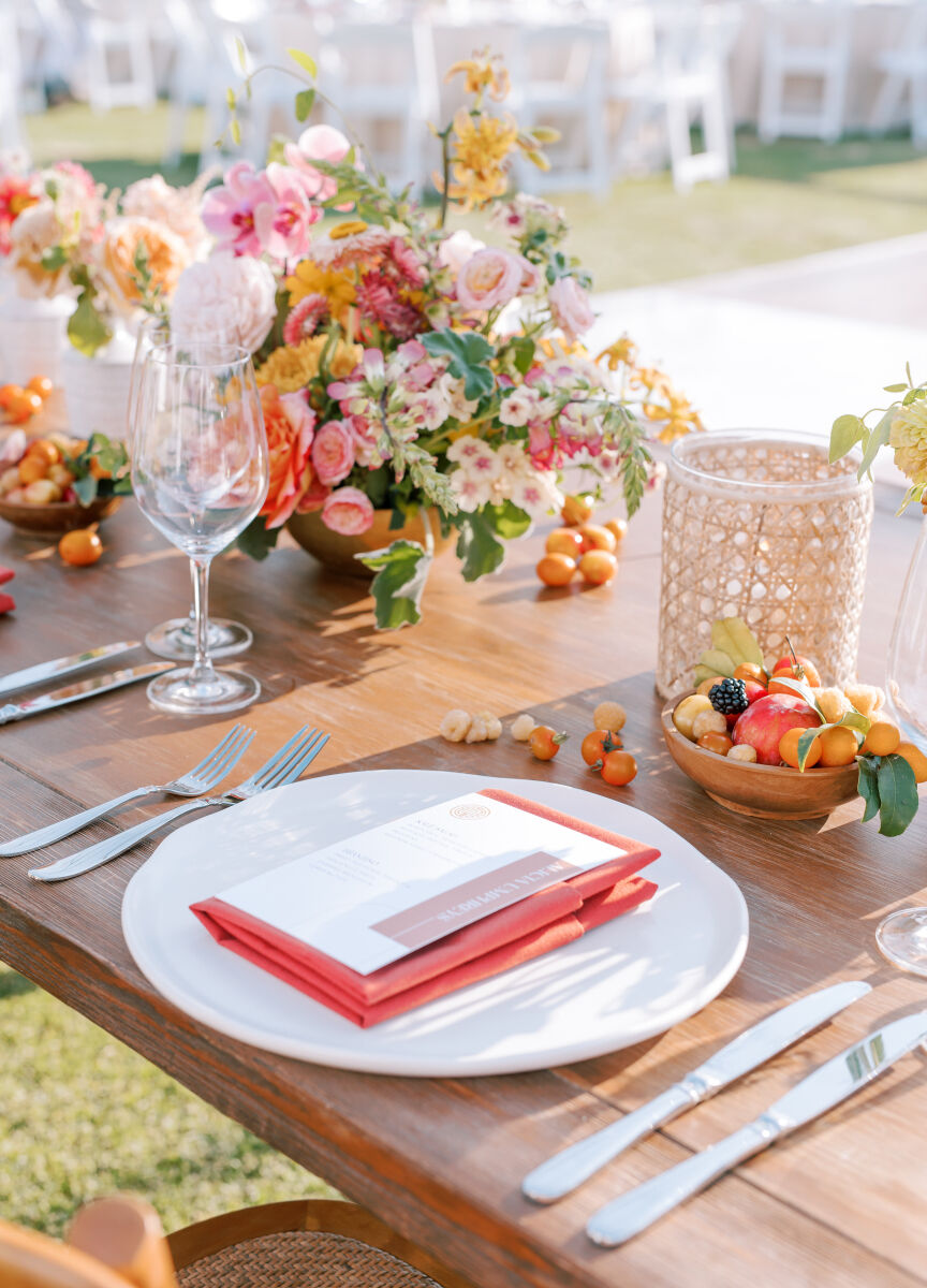 A colorful wedding reception table with coral dinner napkins and centerpieces incorporating both fruit and flowers.