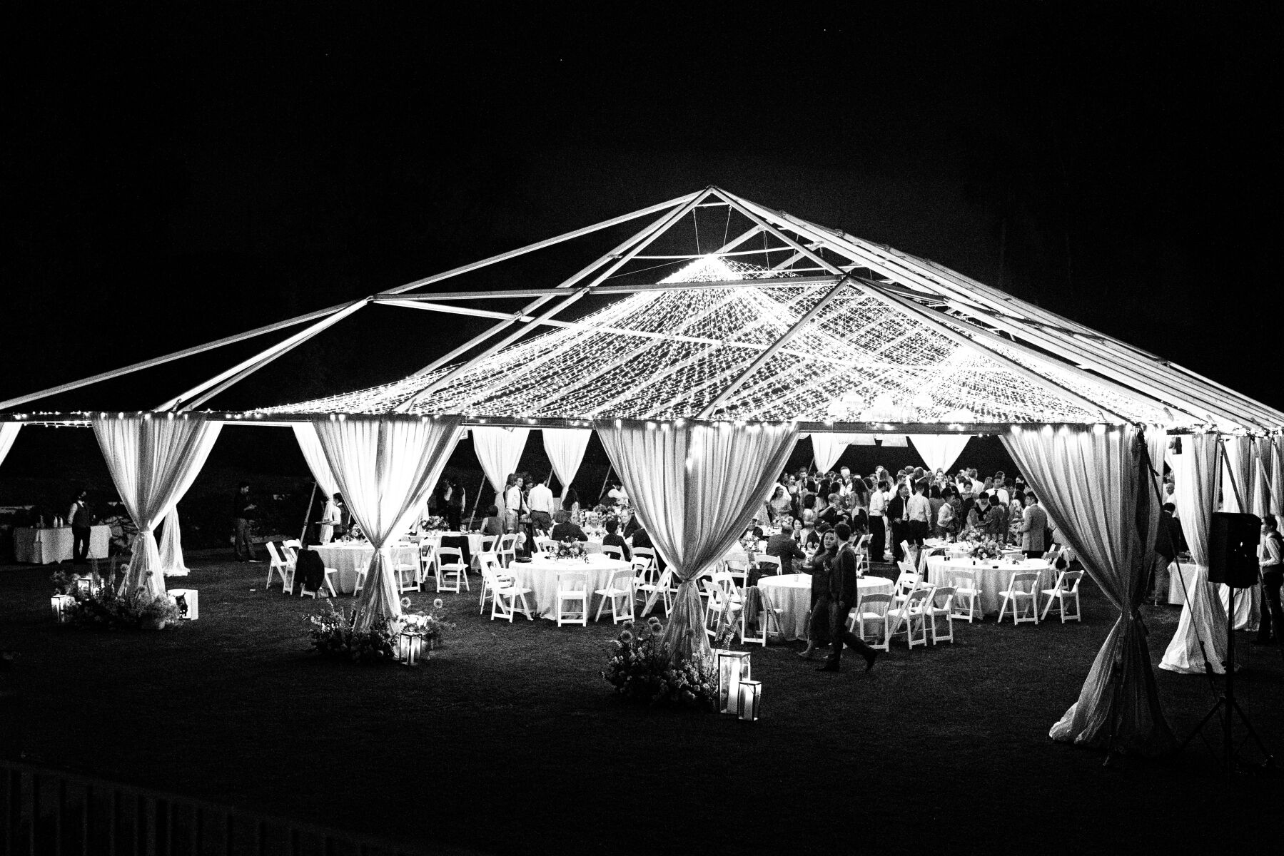 A canopy of twinkly lights illuminates the frame of an open-air tent during an evening wedding reception.